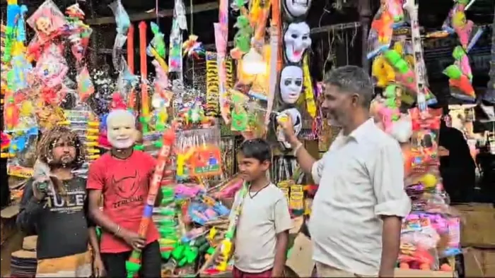 पिचकारी, मुखौटा, गुलाल आदि की खरीदारी करते युवक व बच्चे को कलर फौग उड़ा कर दिखाते दुकानदार